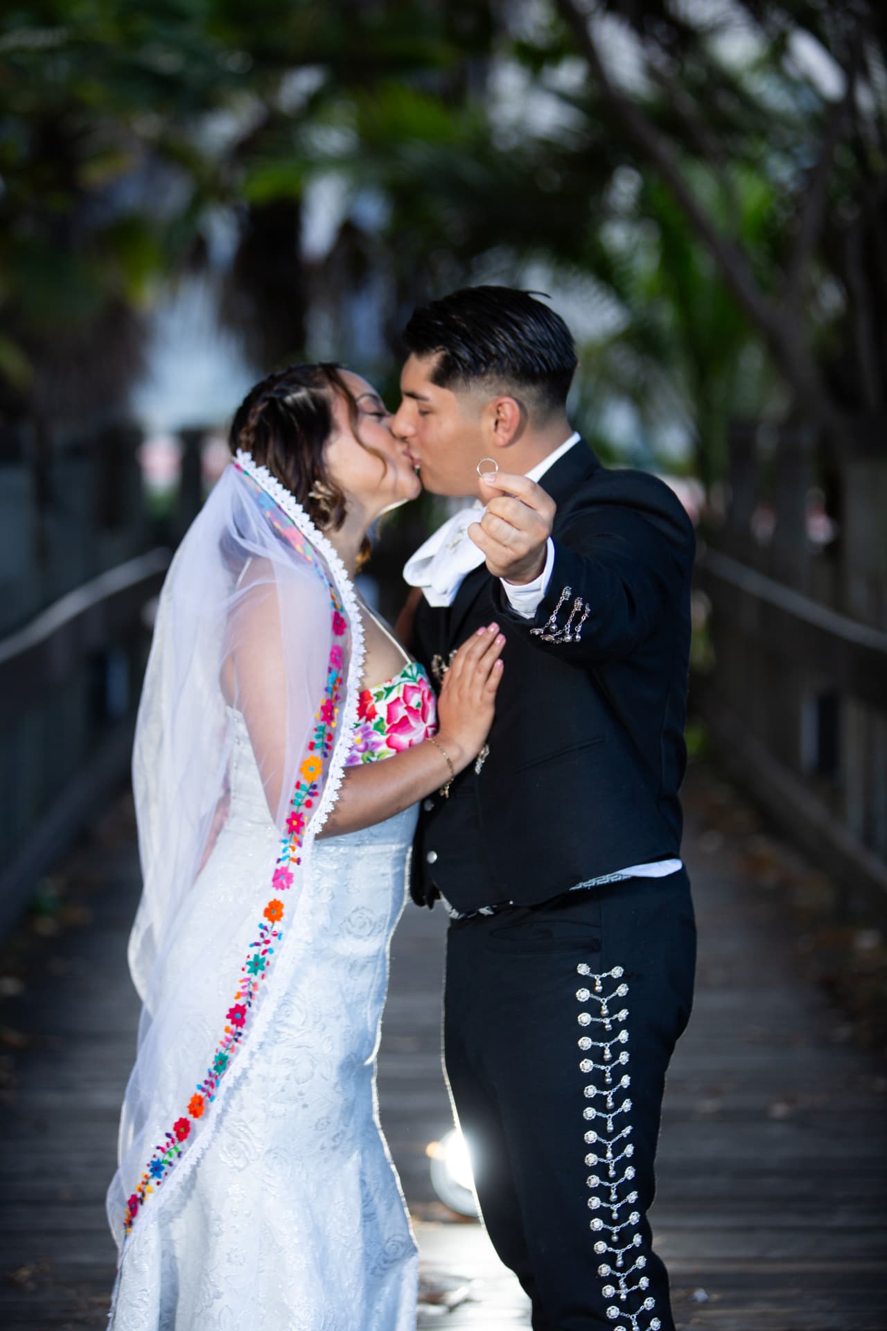 Bride in an embroidered veil and groom in a charro suit share a kiss on a garden bridge
