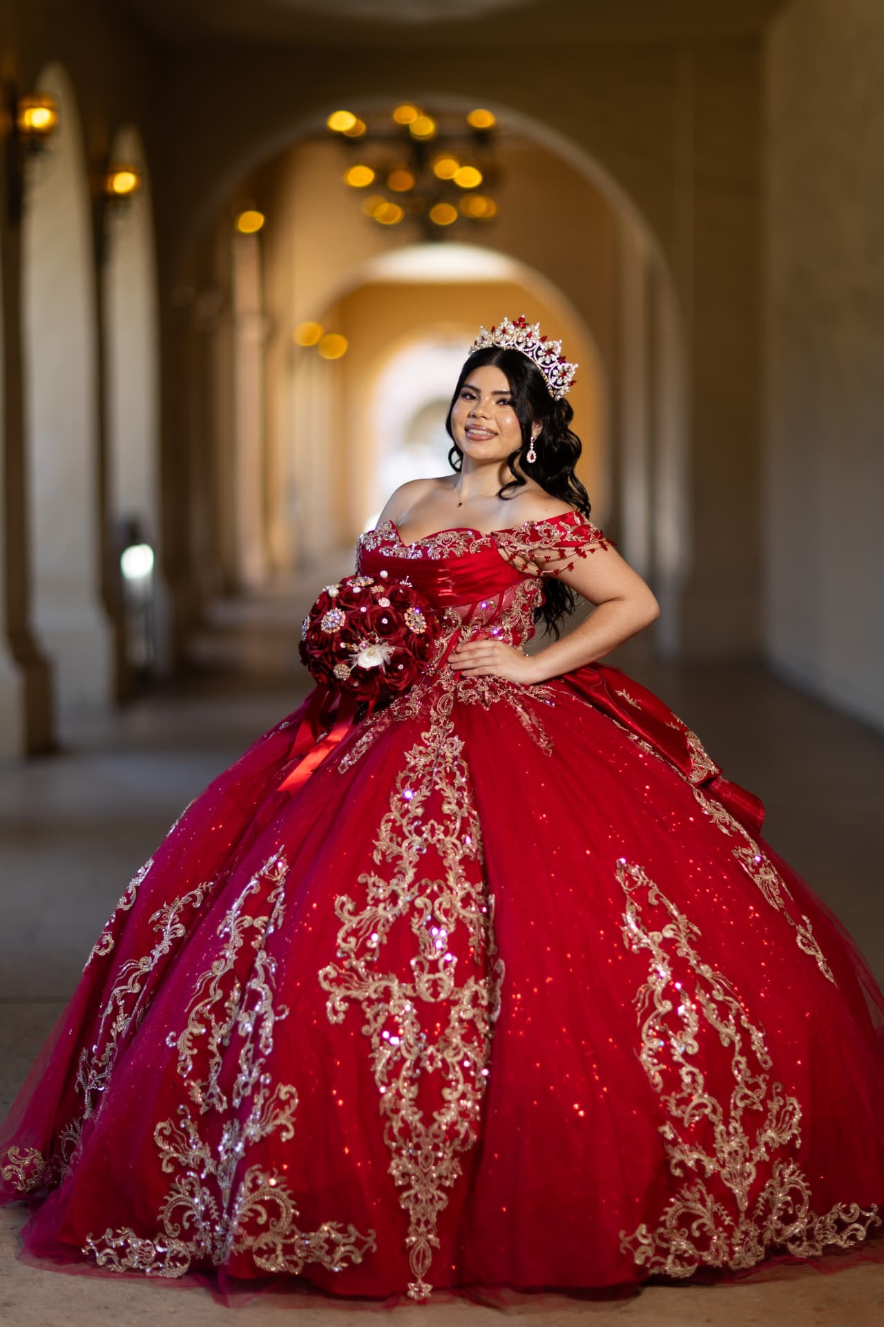 Quinceañera in a red and gold gown smiling in a lantern-lit corridor of arches