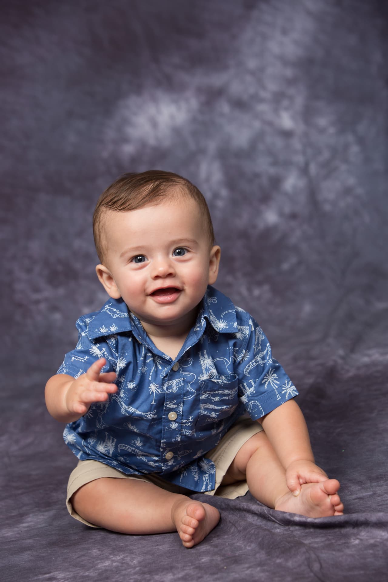 Smiling baby boy seated in a blue patterned shirt against a deep blue backdrop