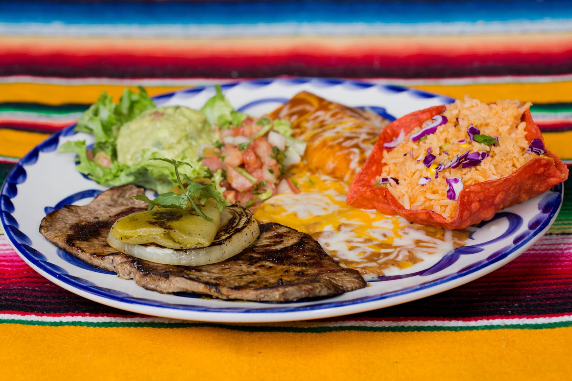 Grilled steak with peppers and onions served with an enchilada, rice, beans, and guacamole