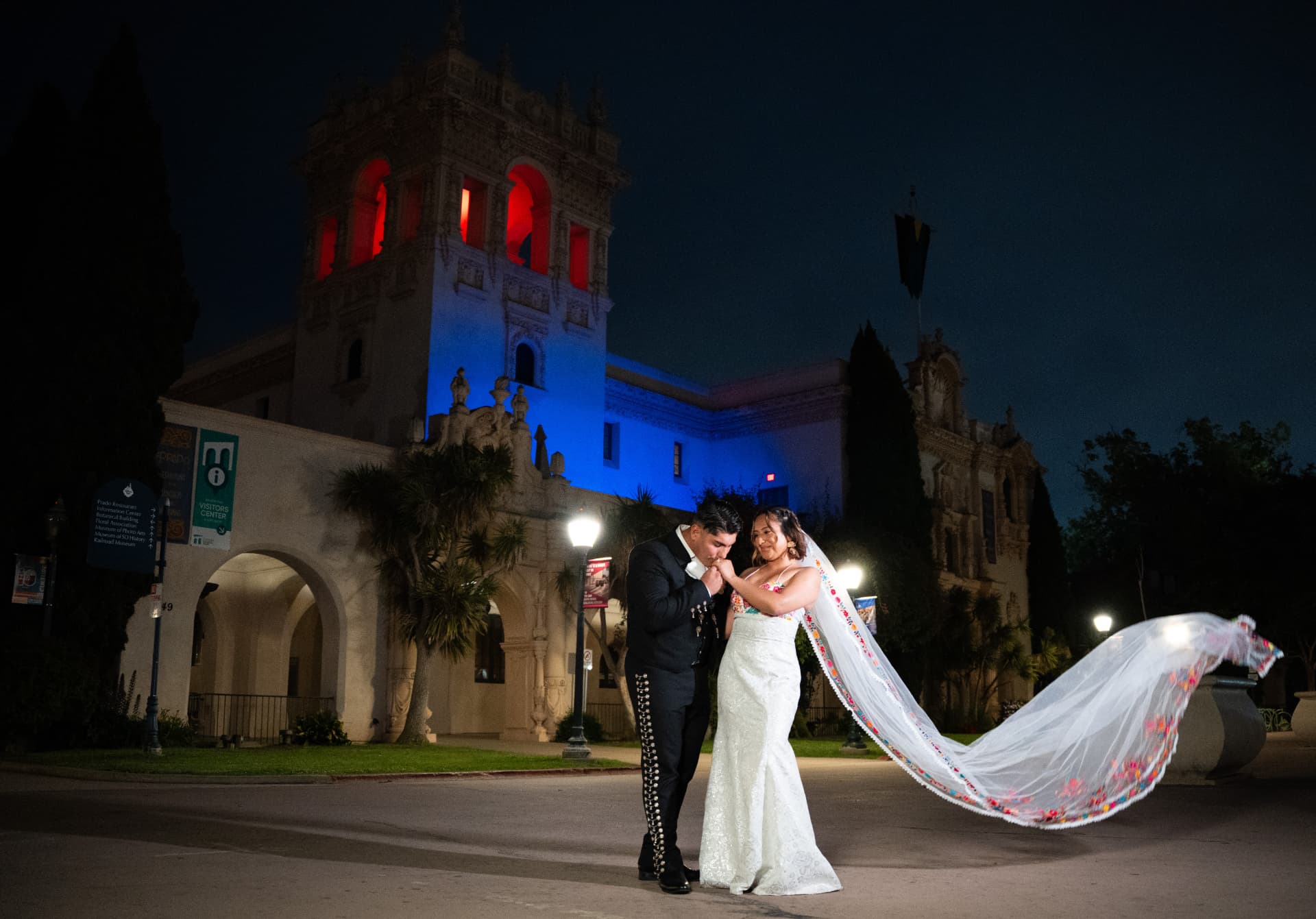 Groom in a charro suit kissing his bride’s hand as her embroidered veil flows at night