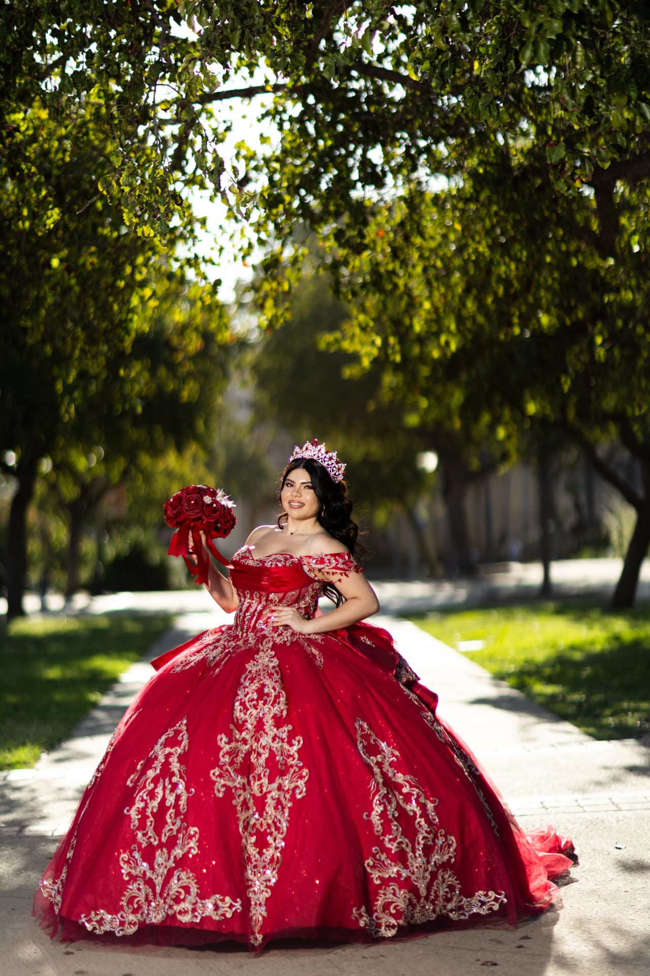 Quinceañera in a red gown raising her rose bouquet on a sunlit garden pathway