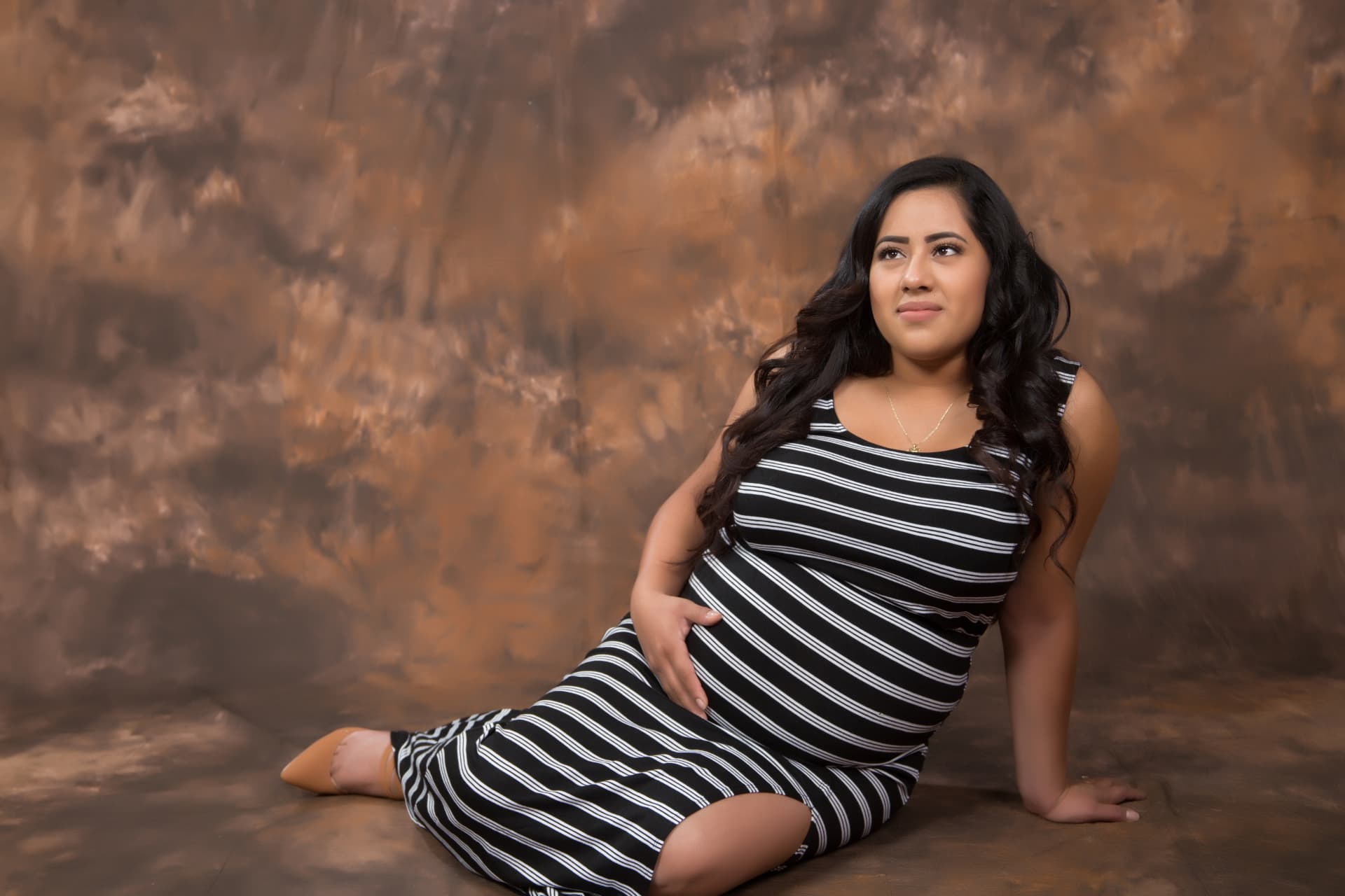Expectant mother seated in a striped dress before a warm studio backdrop