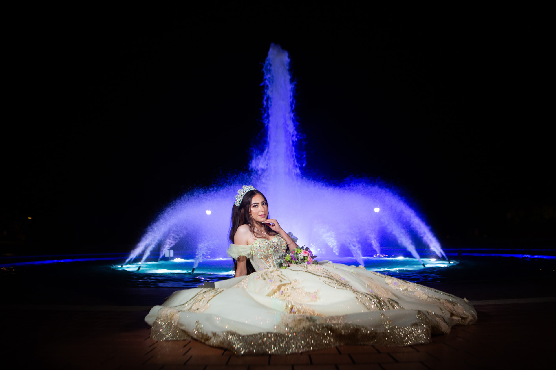 Quinceañera seated in her gown before a blue-illuminated fountain at night