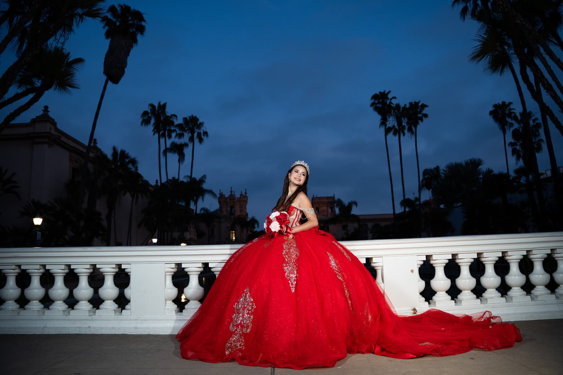 Quinceañera in a red gown seated on a balustrade beneath palm trees at dusk