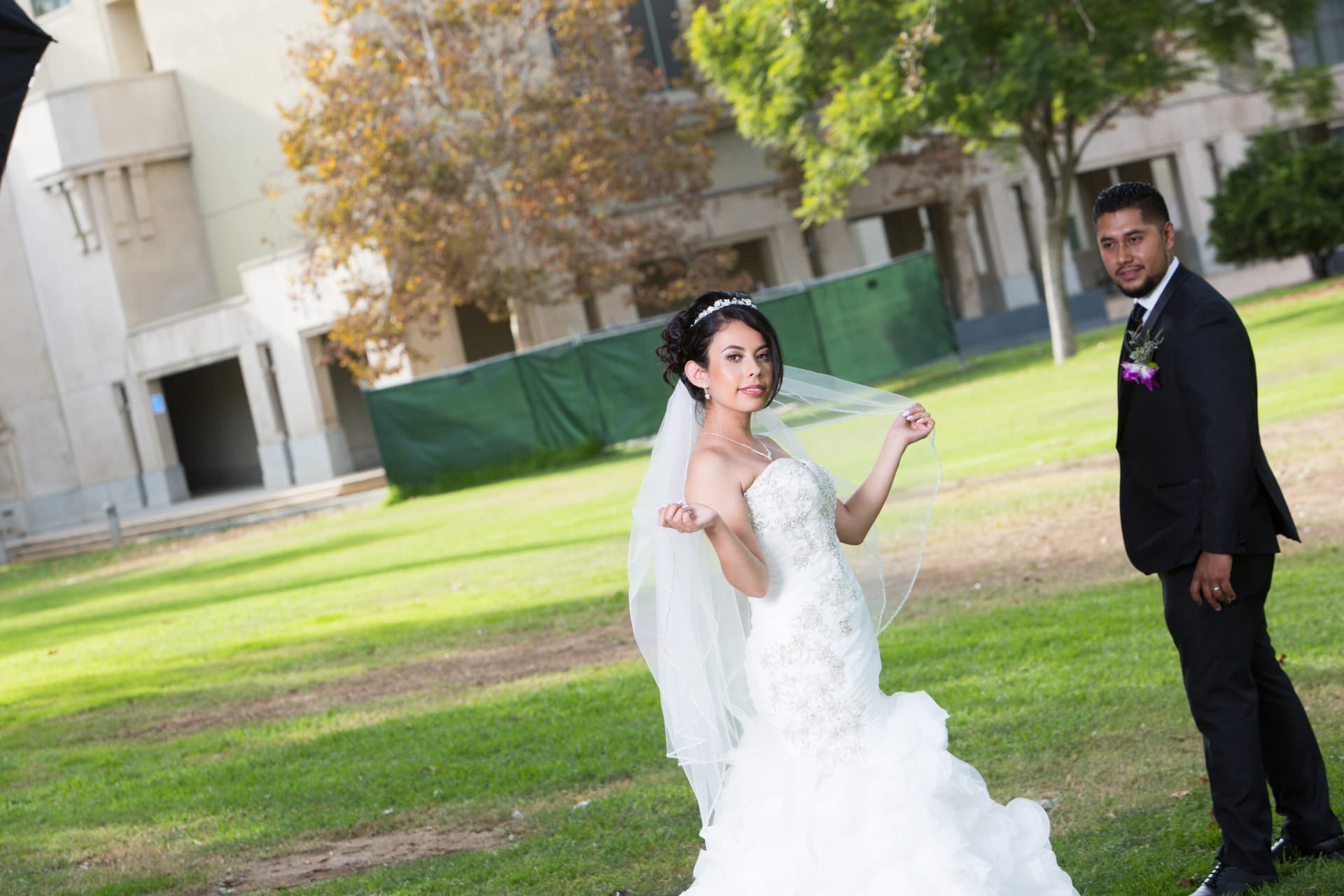 Bride holding her veil in a garden as the groom watches from nearby