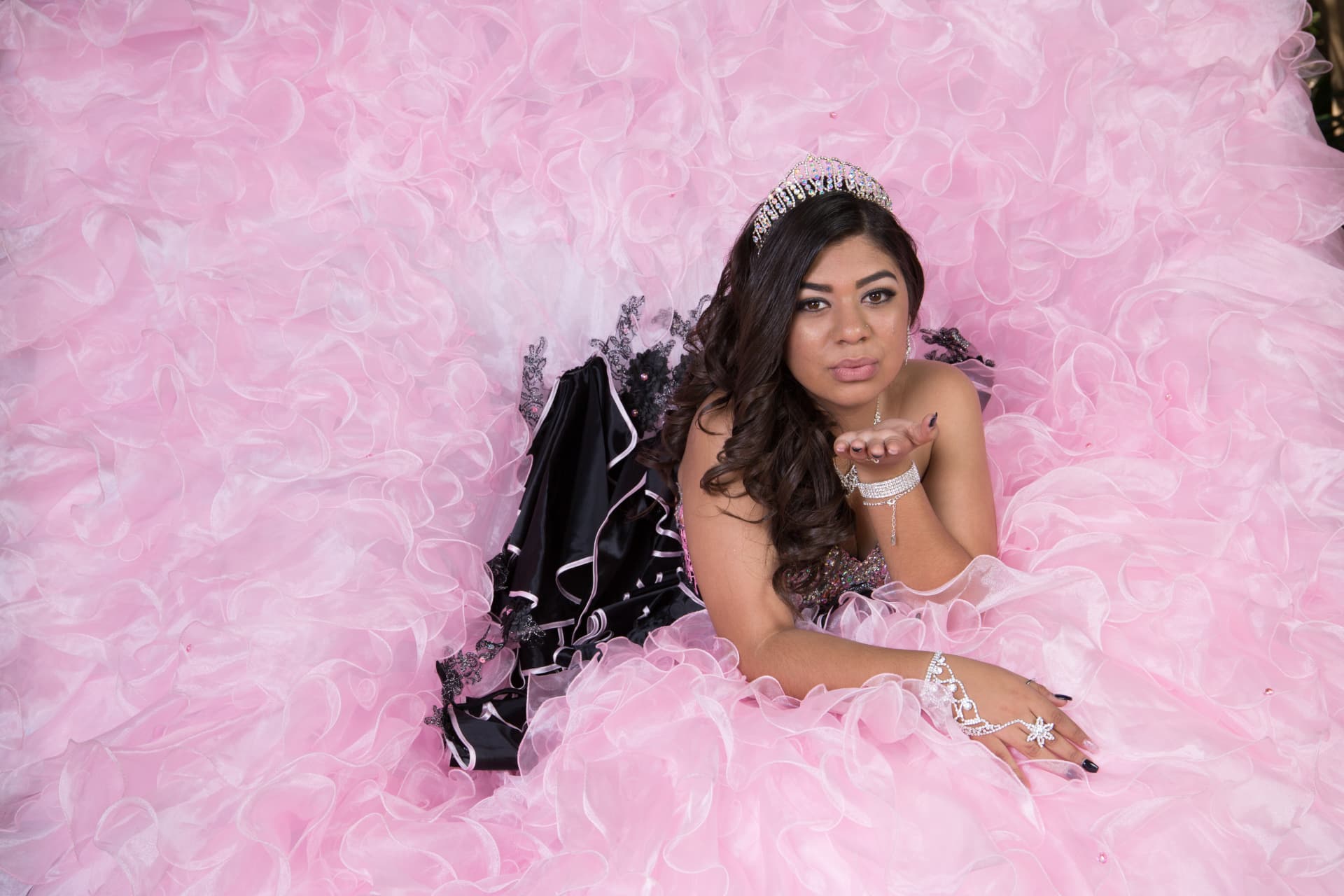 Quinceañera in a pink ruffled gown with black accents reclining and blowing a kiss